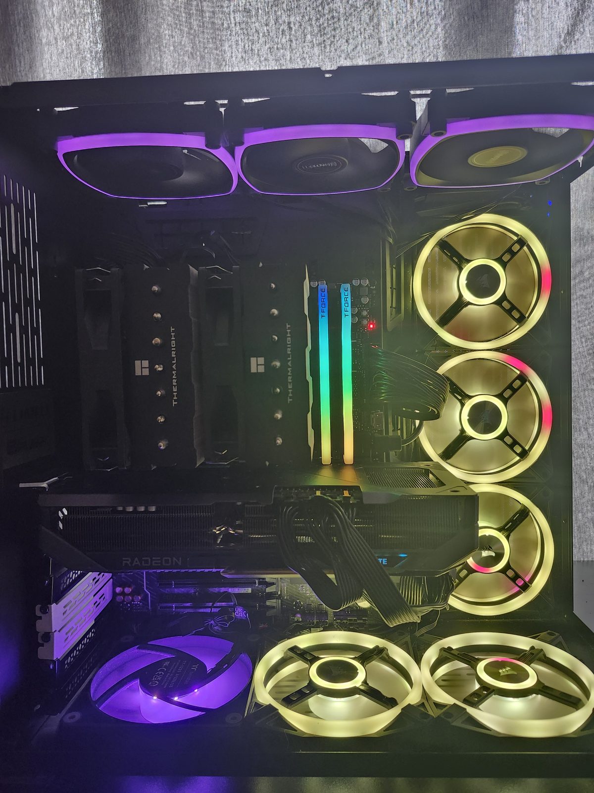 Interior of a dual-chamber PC with the lights on. Purple fans sit up top, yellow and pink fans line the side and bottom, two T-Force RGB memory sticks are lit, and a dual-tower Thermalright cooler covers the CPU.